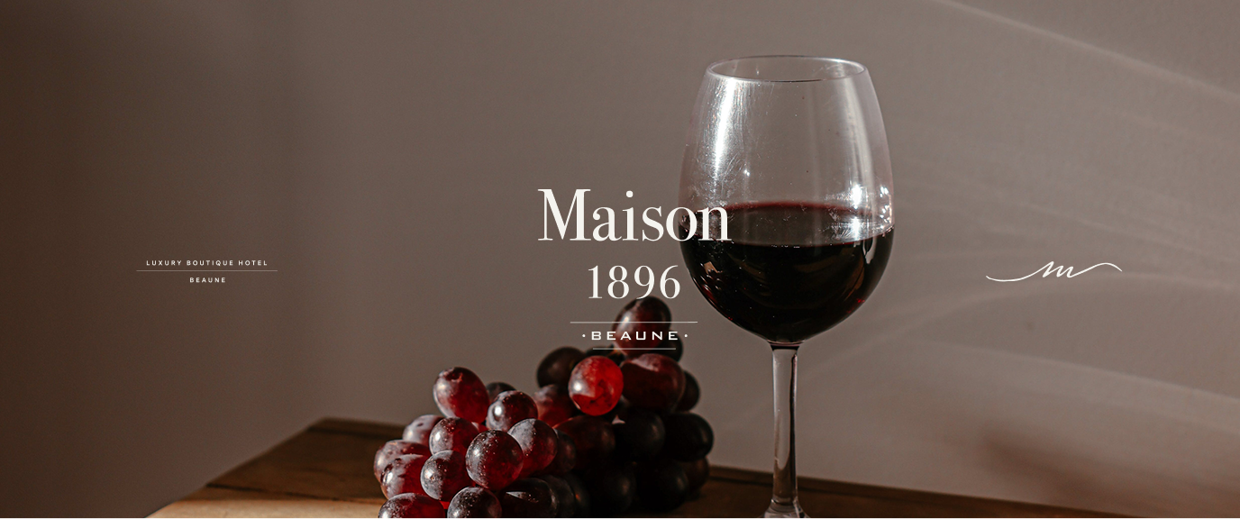 Maison 1896 wordmark, monogram and script signature over a still life with wine glass and grapes